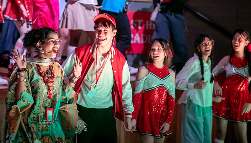 Students performing as Coach Bolton and Mrs Darbus sing near the audience with fellow cast members on stage behind them.